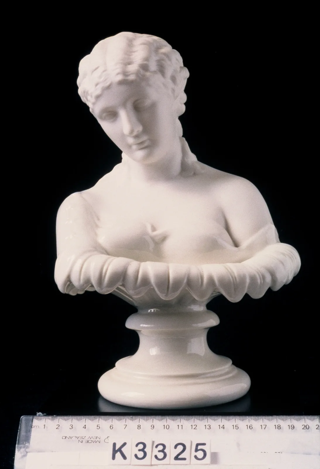 White unglazed porcelain bust of a woman on a turned socle base, photographed against black beside a ruler and a museum reference card reading K3325