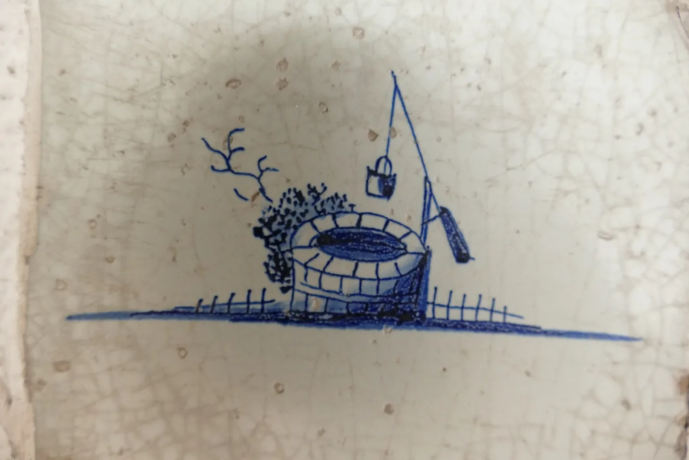 Single blue-and-white Delft tile set into a rough plaster wall, painted with a well and its winch, a bucket on a rope and a low fence, the whole glaze covered in a fine network of crazing
