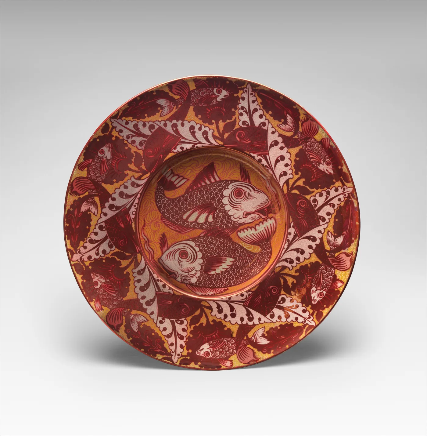 Circular earthenware charger in coppery ruby lustre, its well filled with two large scaled fish and its broad rim with smaller fish among pointed leaves