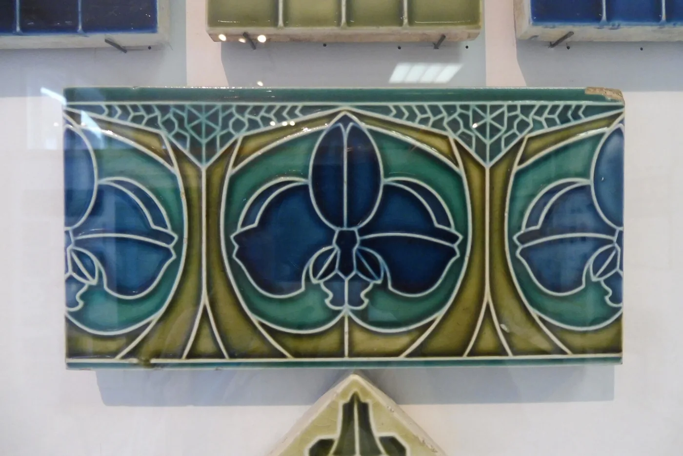 Rectangular Art Nouveau tile in a museum case, decorated with stylised tulip-like blooms in blue, green and olive glazes, each colour area bounded by a raised pale slip line