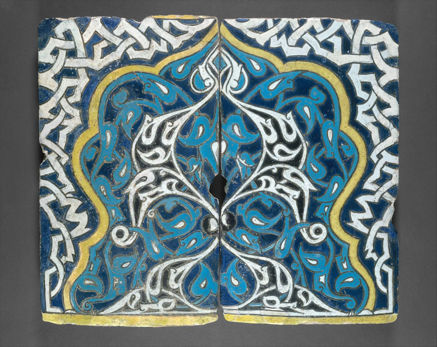 Two shaped tile fragments set together to form one pointed-arch design of white and turquoise arabesques on cobalt, framed by a white interlace band and a yellow line, every colour edged with a narrow dark seam