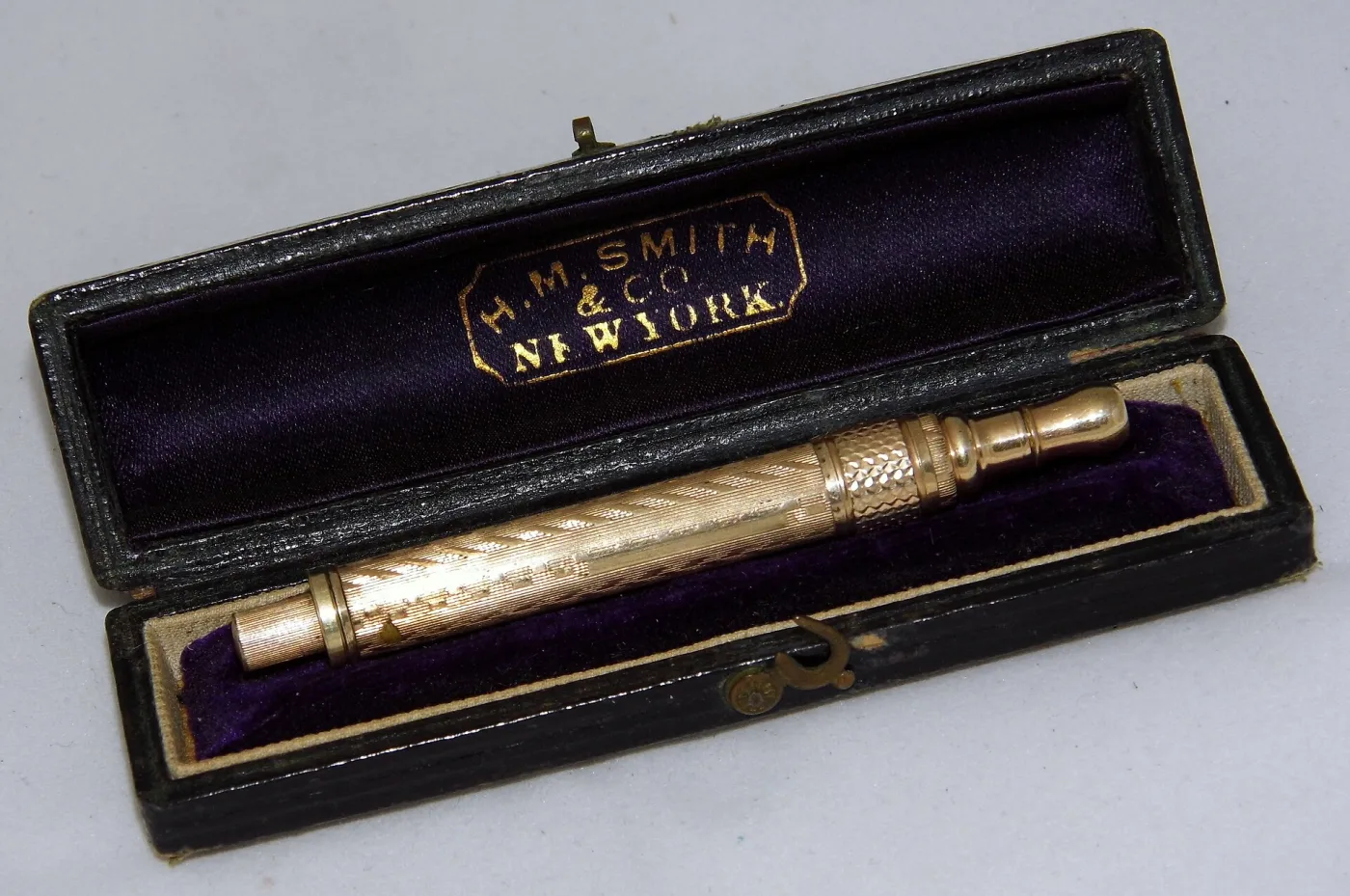 Slim engine-turned metal writing instrument lying in a fitted case lined with purple velvet and silk, the silk lid stamped in gold within a shaped cartouche reading H. M. Smith & Co, New York