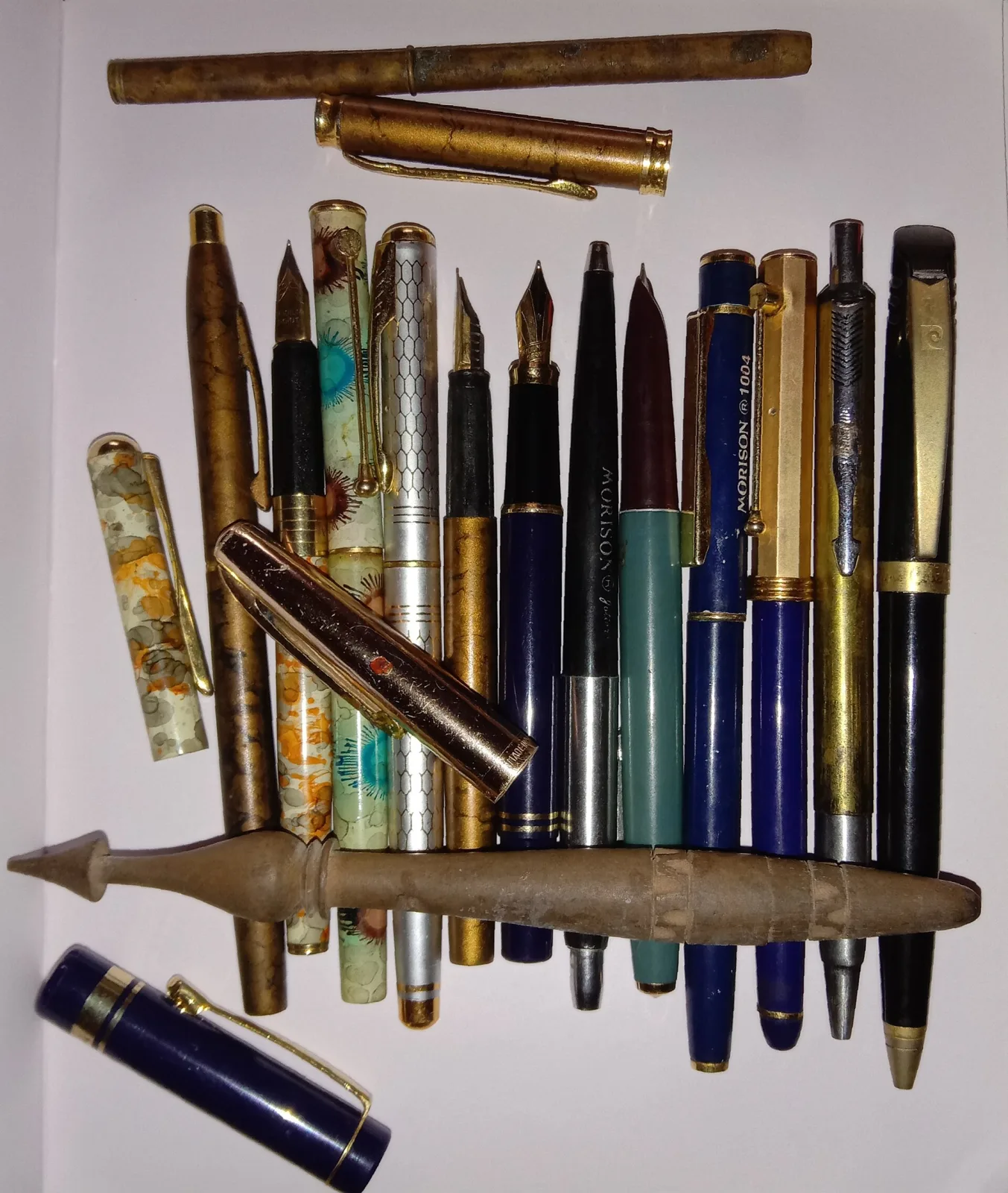 A dozen and a half fountain pens and pencils laid out in rows, in marbled, mottled and plain finishes with visible nibs and clips, arranged around a pointed brass artillery-shell object
