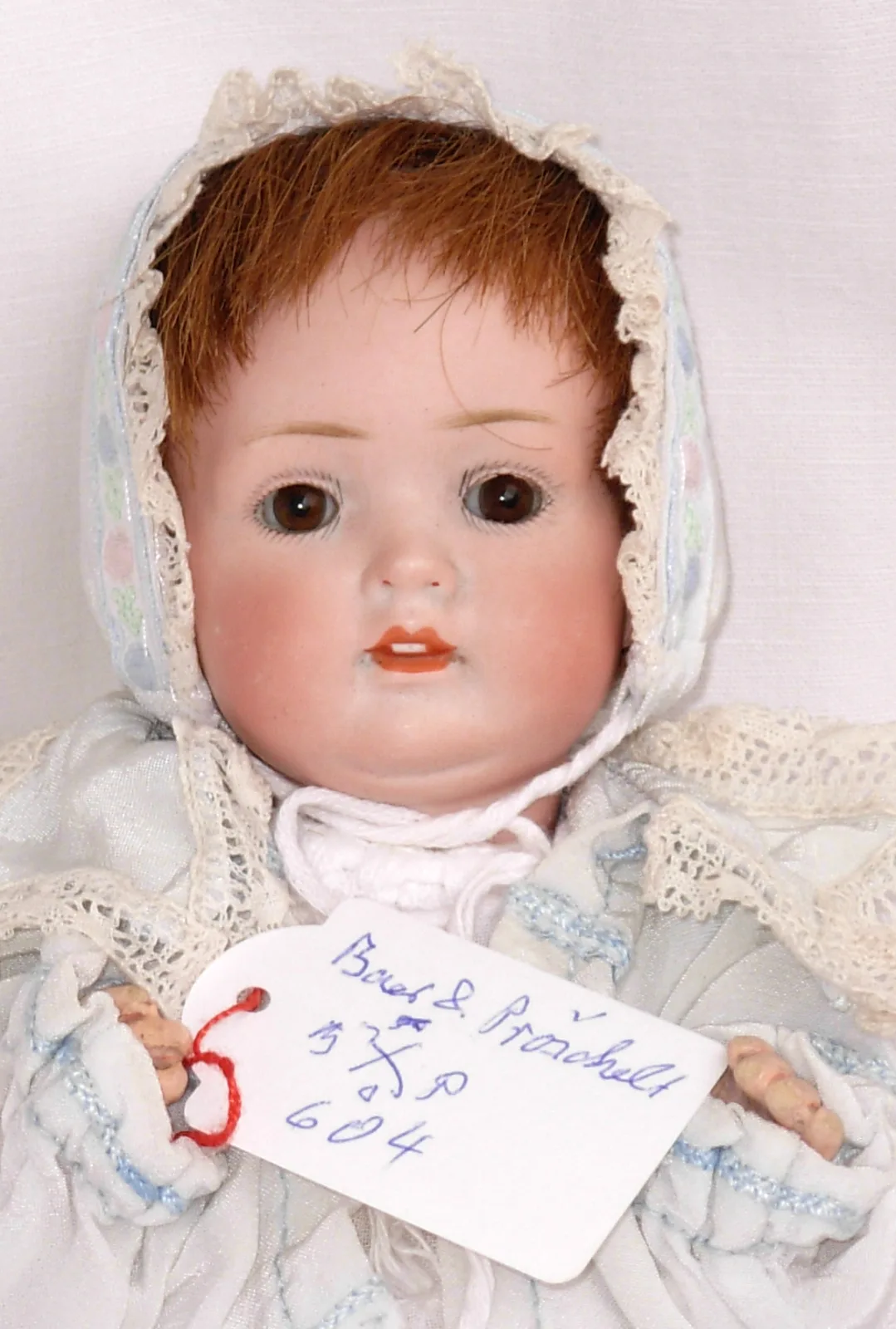 Bisque-head baby doll in a white lace bonnet and gown with a red-haired wig, holding a handwritten museum tag reading Bähr und Pröschild and the number 604