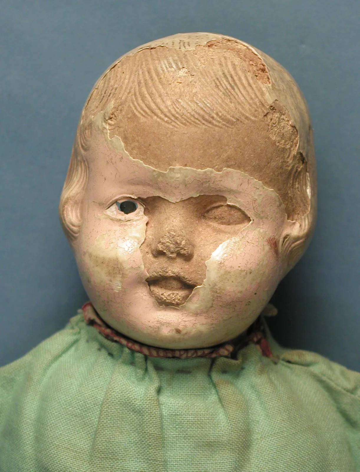 Doll's head of moulded composition with the painted surface crazed into a dense net of fine lines and broken away in large patches, one glass eye missing from its socket, above a green cloth body