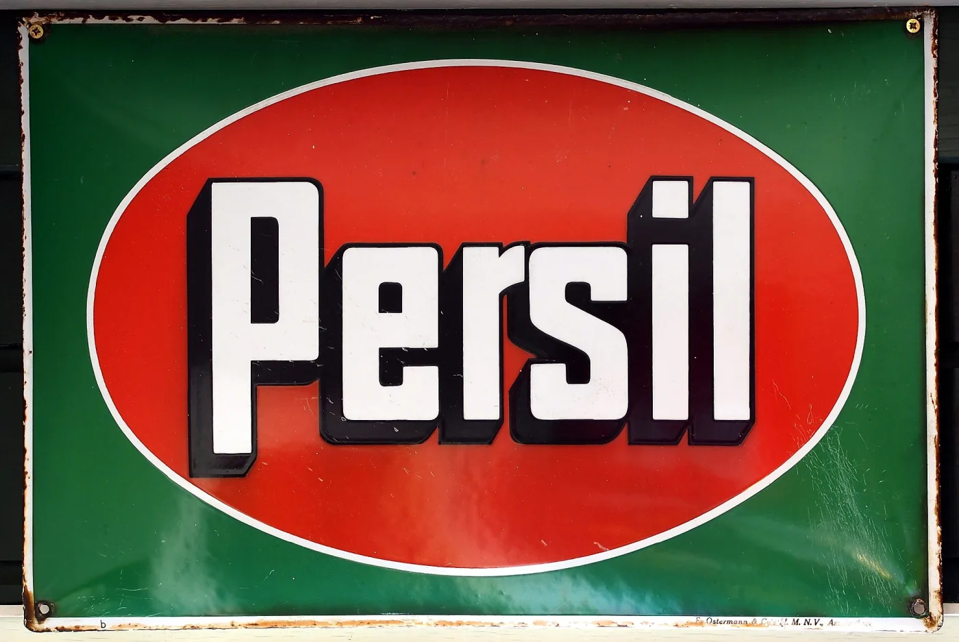 Rectangular enamel sign with a deep green ground carrying a red oval, inside which the word Persil is lettered in white with heavy black shadowing, the sign's edges chipped and its corners pierced