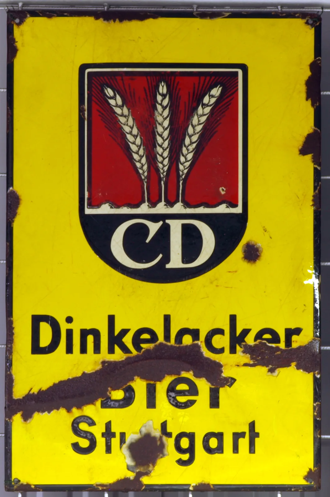 Enamel sign on a yellow ground lettered Dinkelacker Bier Stuttgart above a black shield with wheat ears, the lower third eaten away by rust with the enamel gone and brown staining bleeding upward into the yellow