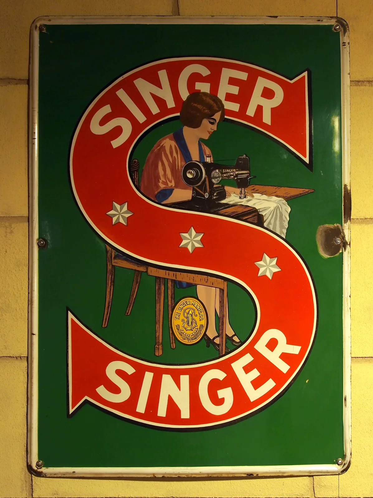 Rectangular enamel sign on a dark green ground with a large red arrow-ended S enclosing the word SINGER twice, and a seated woman at a treadle sewing machine painted inside the letter