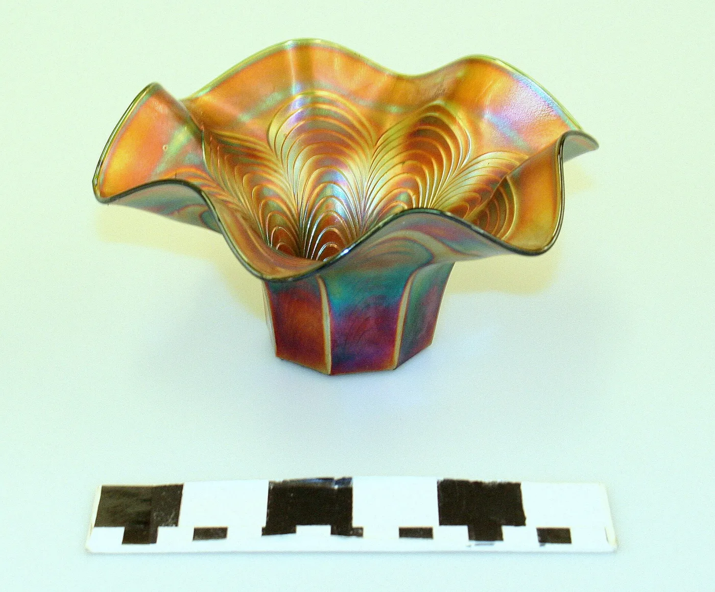 Marigold iridescent glass dish with a deeply fluted upturned rim and a moulded drapery pattern radiating from the centre, photographed above a black-and-white scale bar