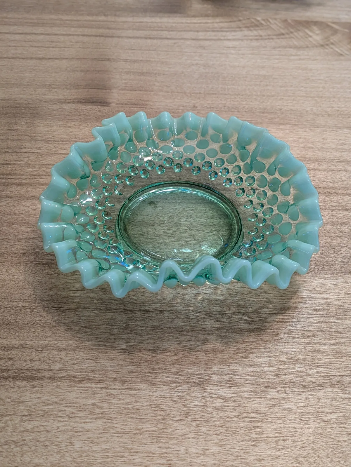 Small opalescent hobnail dish in pale yellow-green, its crimped rim and the tips of the hobnails whitening towards the edge, resting on a wooden surface