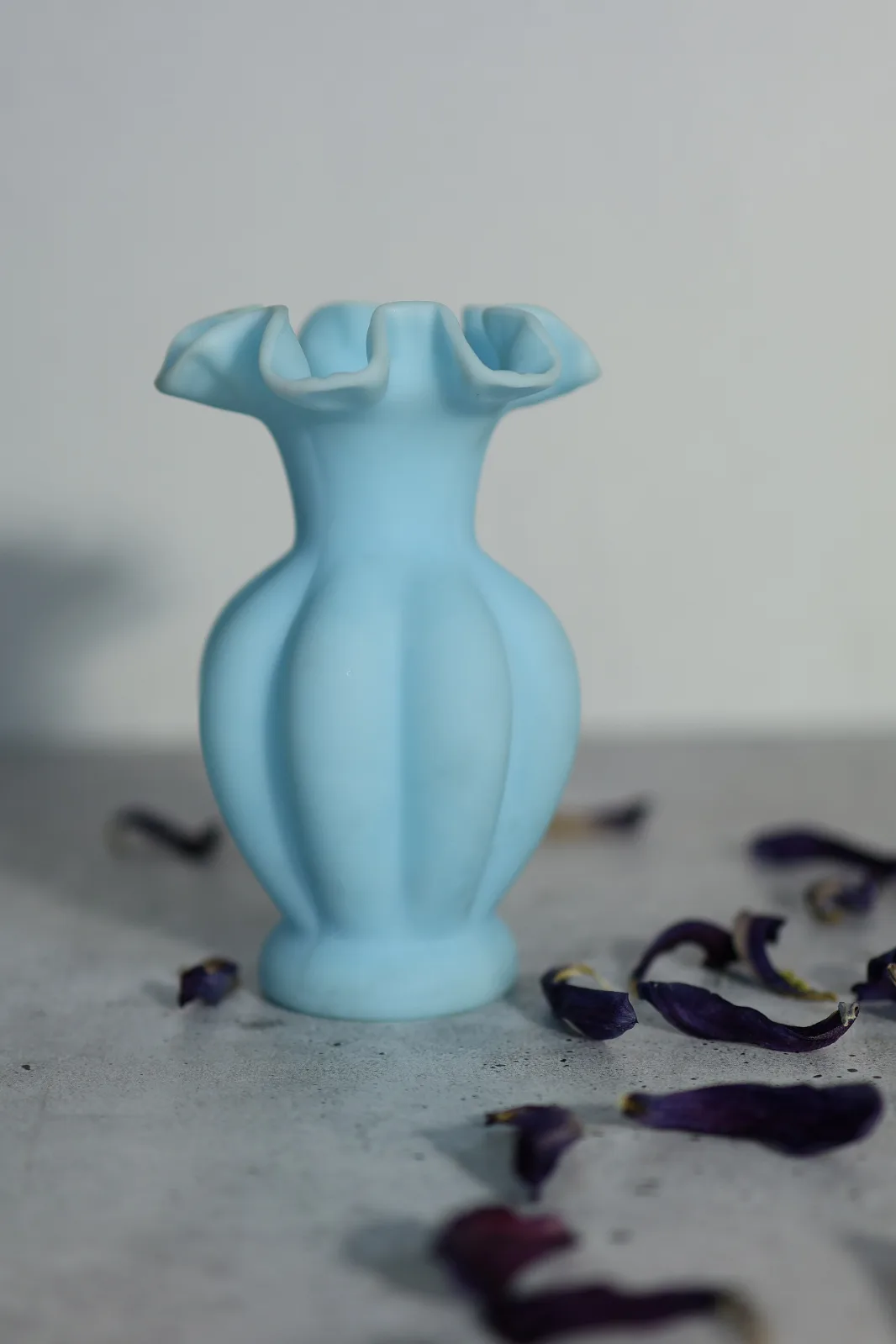 Satin-finished pale blue vase with vertical melon lobes and a crimped rim, on a grey surface scattered with dried petals