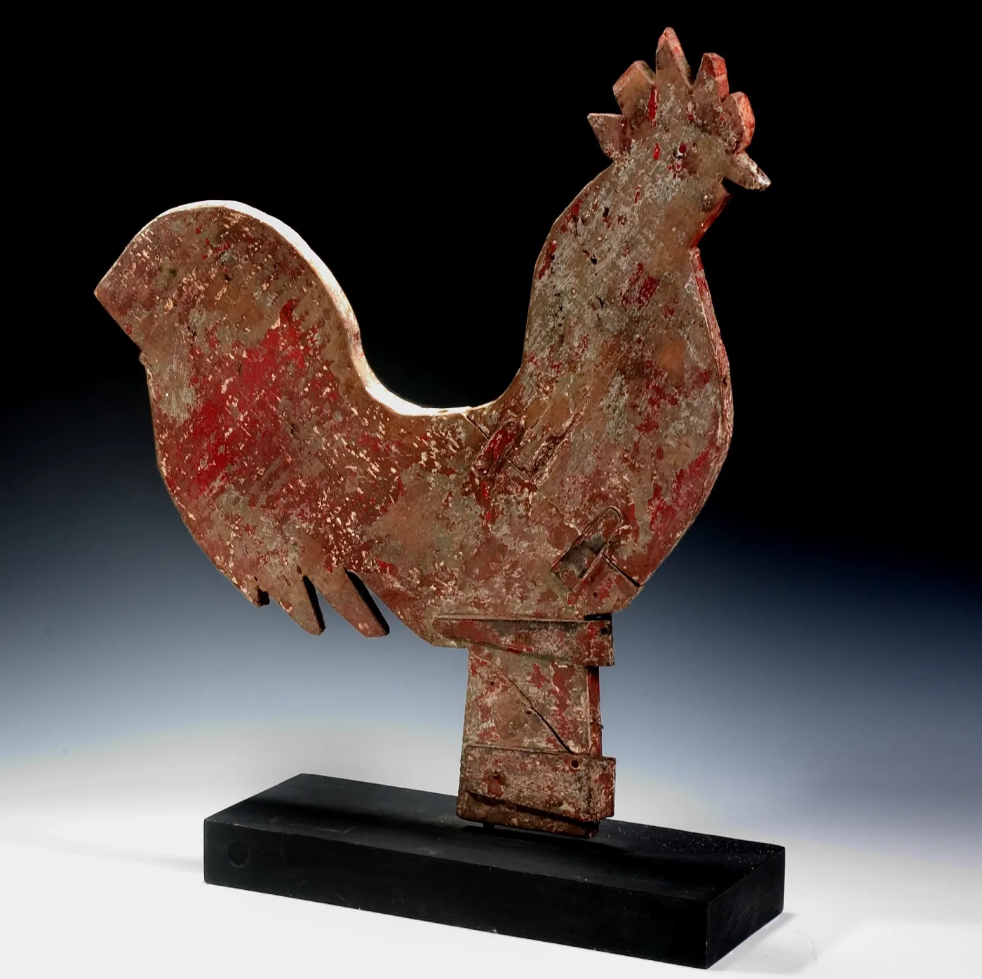 Flat wooden rooster weathervane with worn red paint, sheet-metal patches at the neck and body, mounted on a black display block