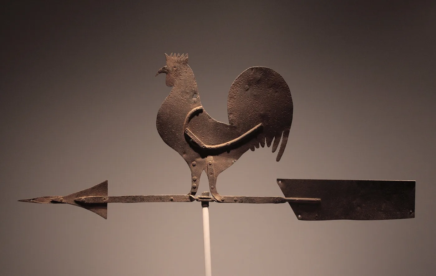 Rusted sheet-iron rooster weathervane with an applied riveted wing, mounted on a horizontal rod with a pointed arrow at one end and a flat rudder at the other