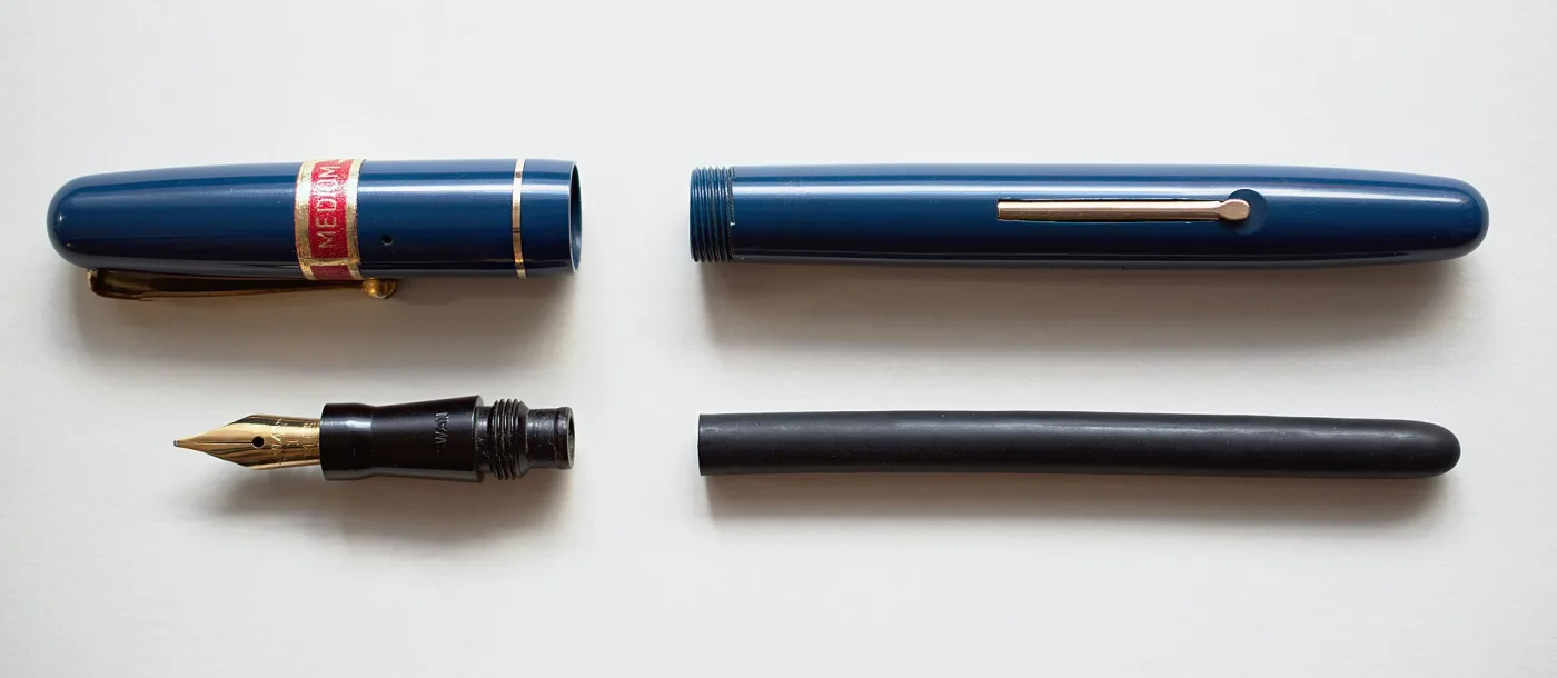 Blue-black fountain pen shown in four pieces on a white ground: the capped upper section with a red band, the separate cap, the barrel, and the section with its gold nib withdrawn