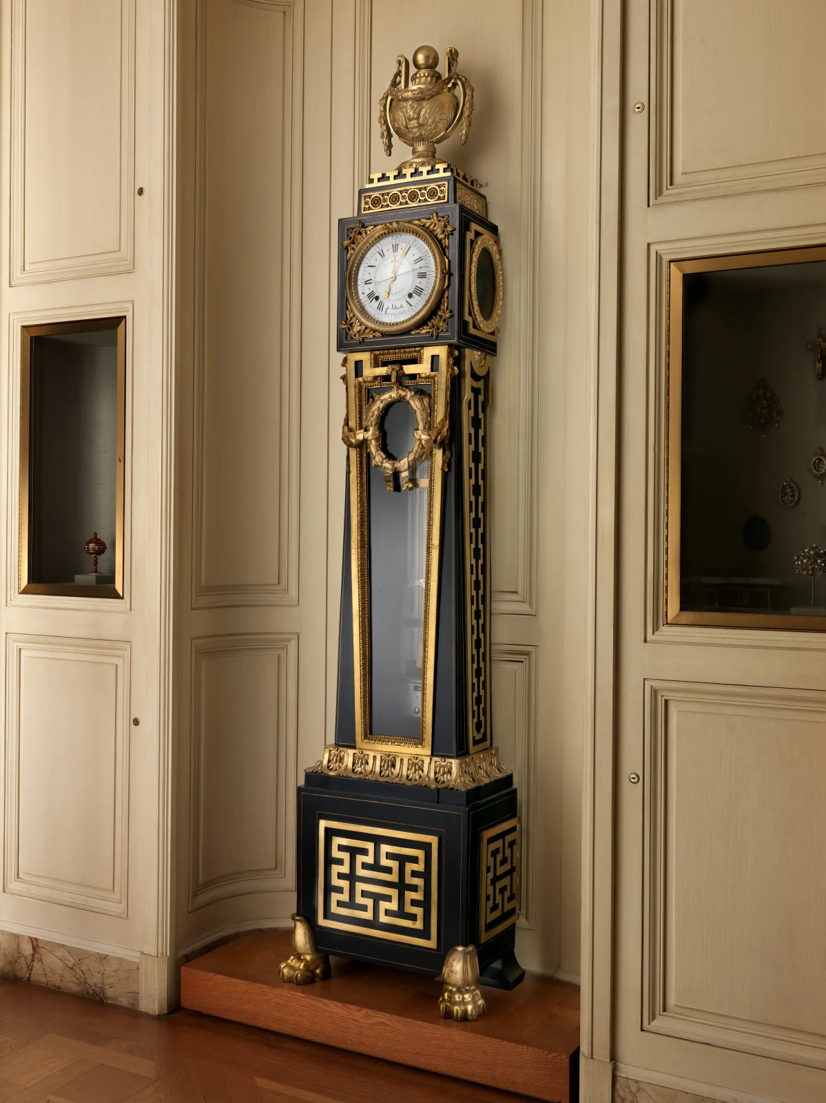 Tall dark clock case with gilt-bronze mounts standing in a panelled interior, its glazed trunk showing a long pendulum, with a white circular dial in a squared hood above a plinth on paw feet