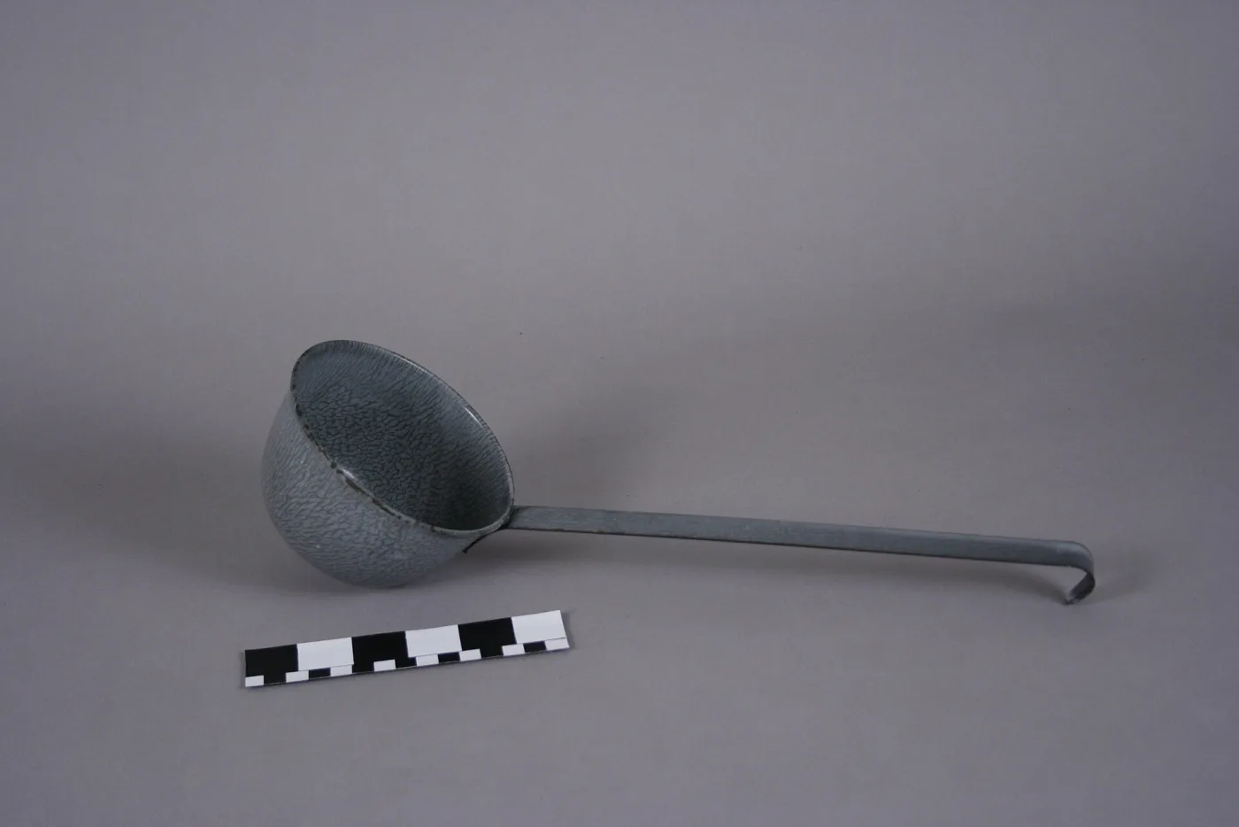 Long-handled dipper with a deep grey enamelled bowl and a plain iron handle hooked at the end, photographed against a grey ground with a scale bar