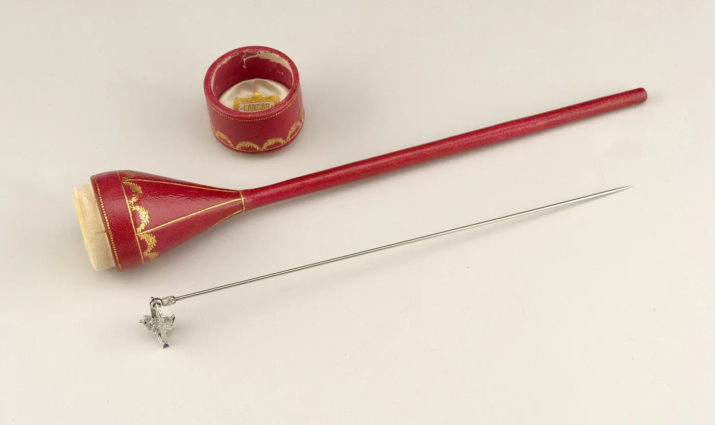 Hat pin with a small gem-set white metal head lying beside its red leather fitted case, the case lid stamped in gold with a retailer's name