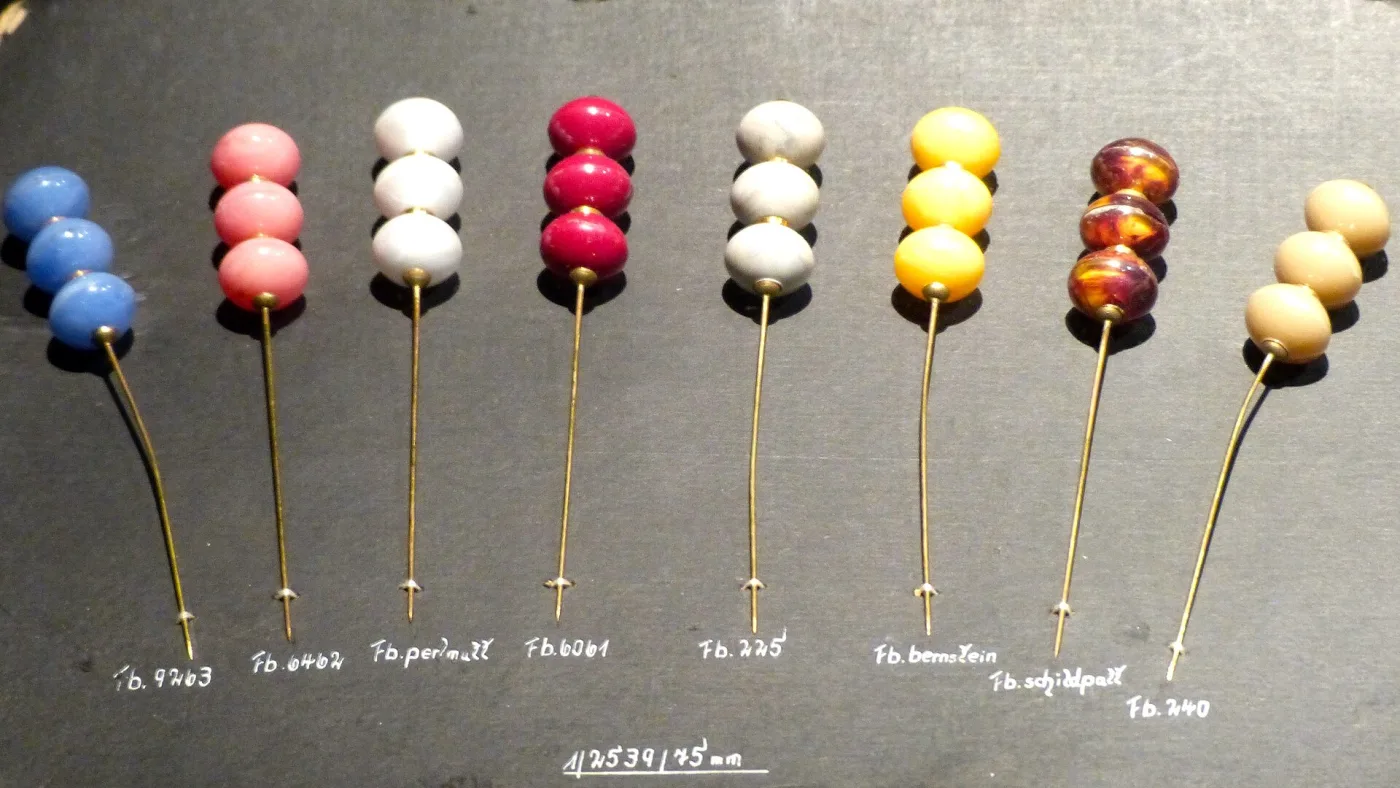 Manufacturer's sample card holding eight hat pins in a row, each with three stacked coloured beads, with hand-lettered colour codes written beneath them