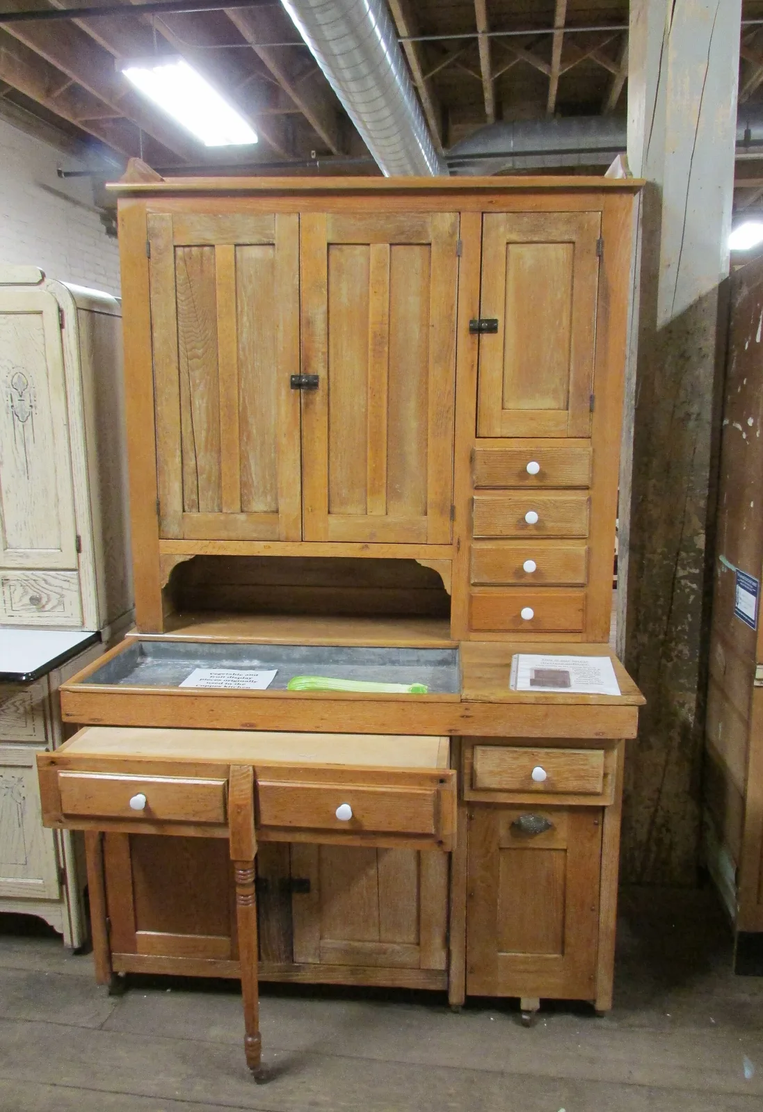 Early kitchen cabinet in pale oak with vertically boarded doors, black cast-iron strap hinges, white porcelain knobs, a zinc-lined open trough work surface and a turned wooden leg supporting a pull-out board