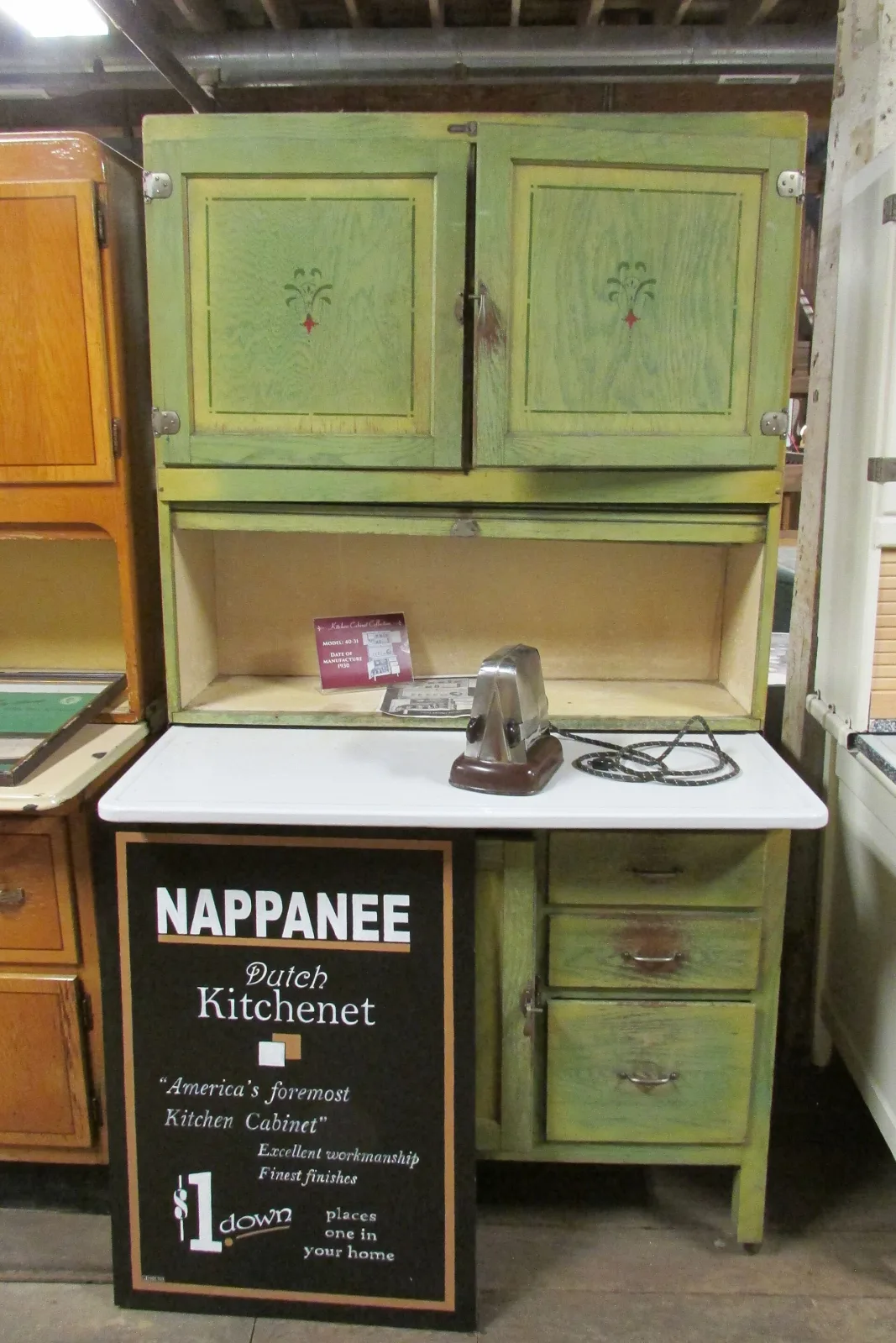 Pale green painted kitchen cabinet with stencilled floral motifs and a red accent on the upper door panels, pressed-steel hinges, stamped drawer pulls and a white enamel pull-out work surface