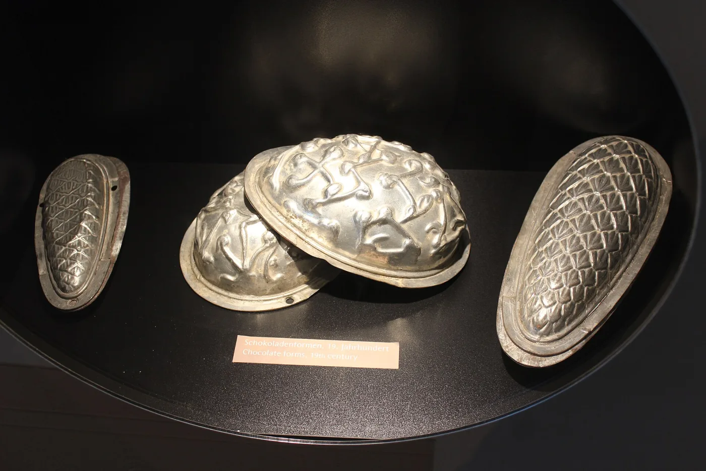 Four two-part metal chocolate moulds displayed in a museum case, formed as textured egg and pine-cone shapes with wide flanged rims
