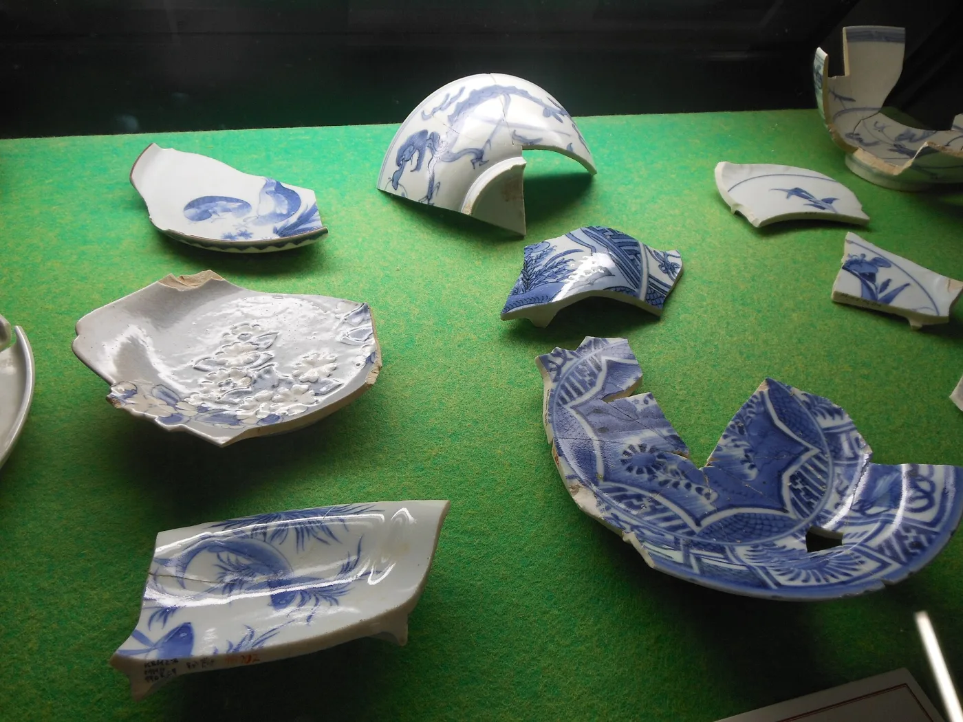 Broken fragments of blue-and-white porcelain laid out on a green cloth, including bowl rims, footrings and body sherds, several showing the thickness of the body in section at the break
