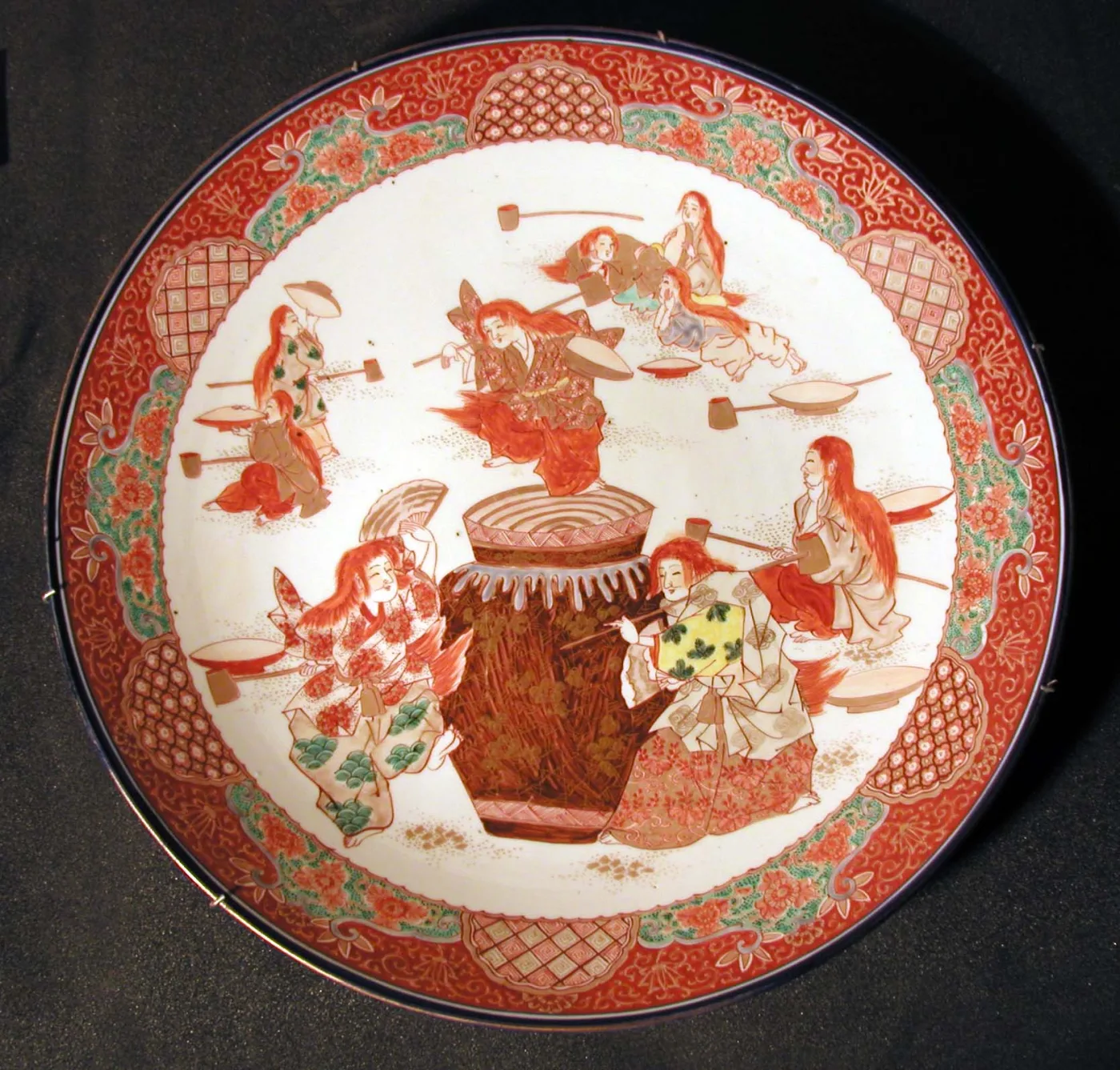 Face of a large Imari charger painted with a scene of figures gathered around a drum, framed by an iron-red and gold rim divided into panels