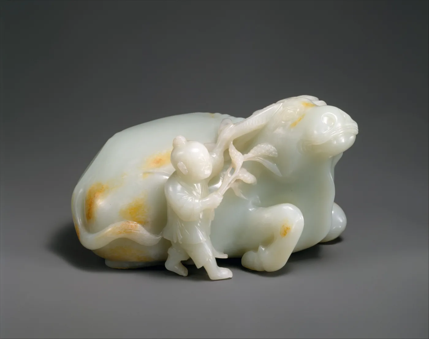 Pale white jade group of a reclining water buffalo with a boy standing at its shoulder, patches of orange staining across the flank