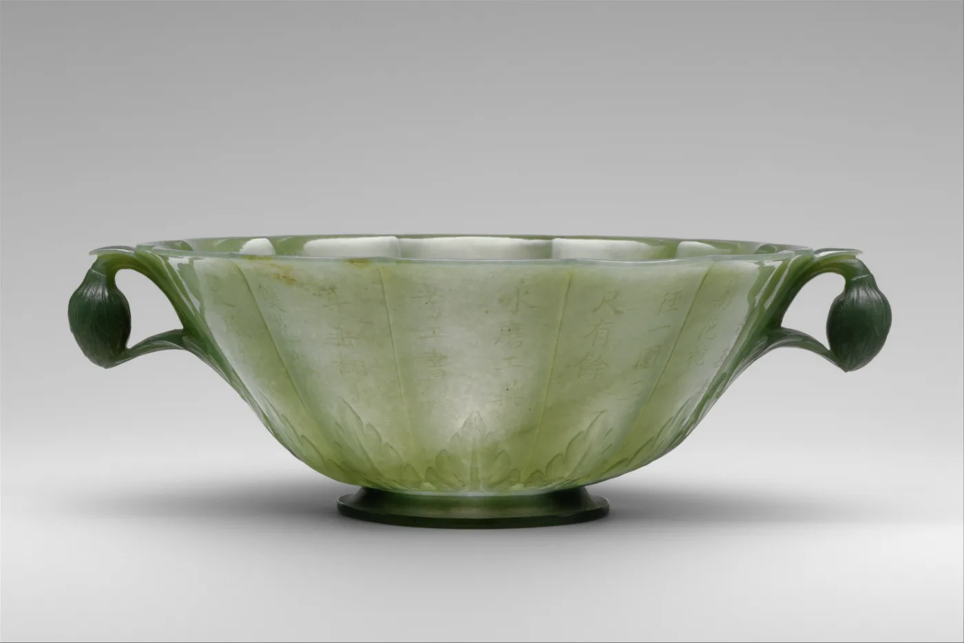 Shallow pale green jade bowl with fluted petal-shaped sides and two dark leaf-and-bud handles, columns of Chinese characters incised across the outer wall