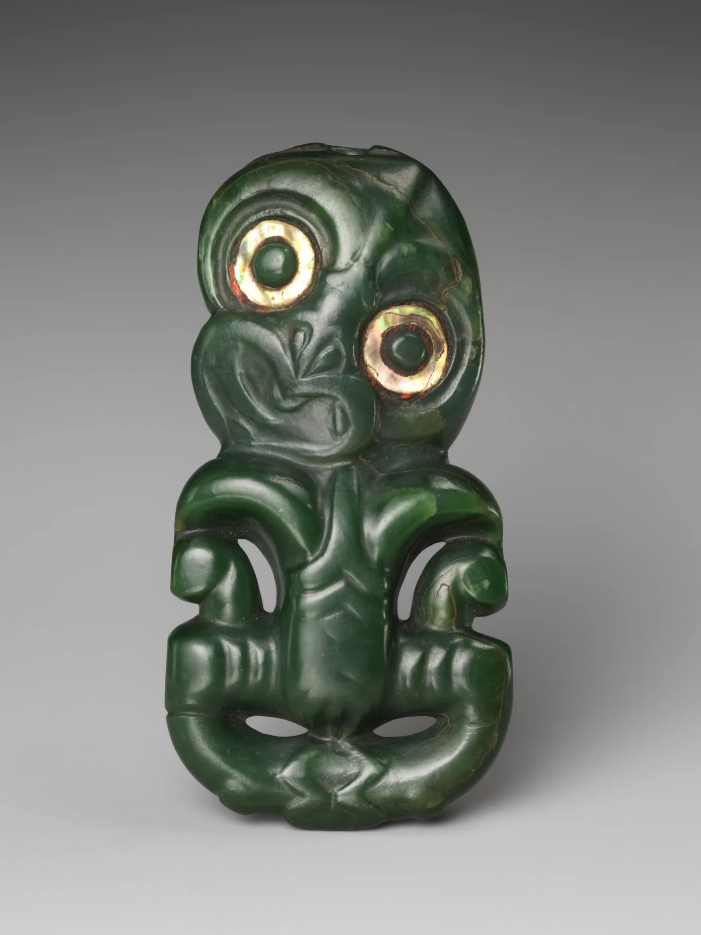 Dark green pendant of a squat figure with a tilted head, hands on thighs, and two large eyes ringed with iridescent shell