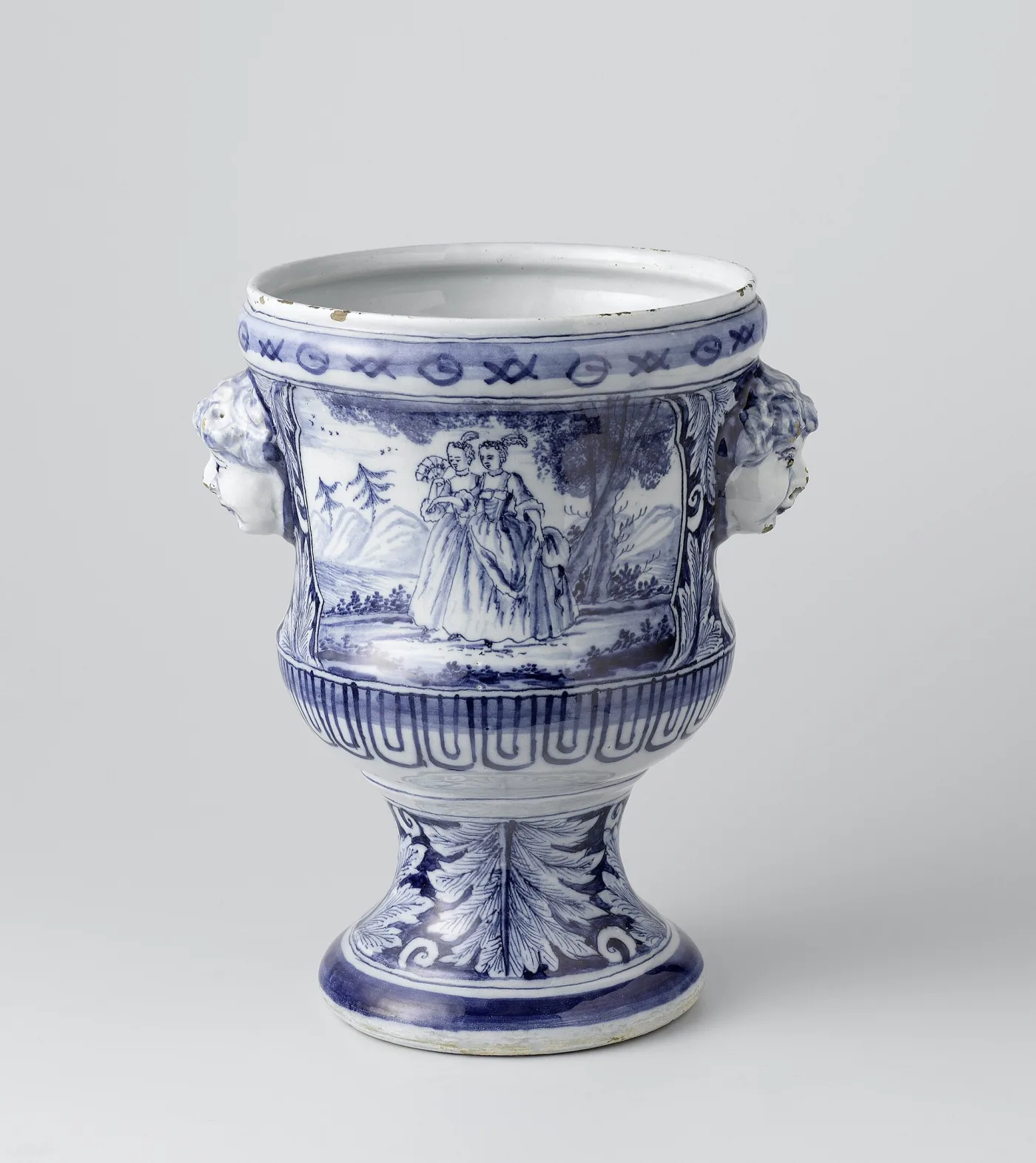 Delft blue and white cache pot on an acanthus moulded pedestal foot, with handles modelled as children heads, painted with two figures walking in a landscape