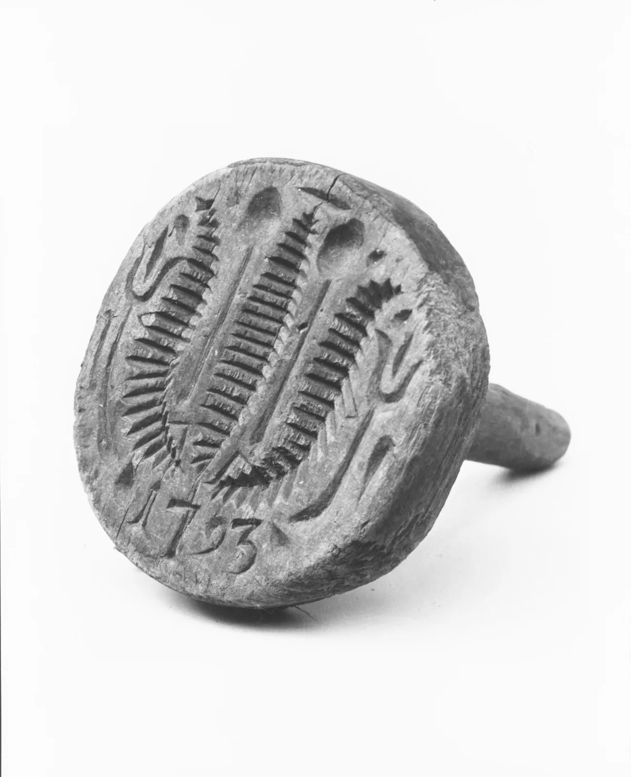 Round wooden butter mould photographed in black and white, its face deeply carved with wheat sheaves and hearts around the incised date 1793, with a turned handle behind