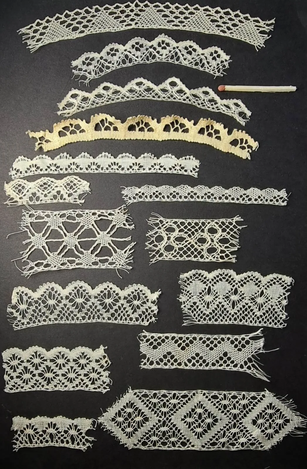 Card mounted with about a dozen short lengths of bobbin lace of different widths and patterns arranged in rows against a black ground, with a lace bobbin laid beside them