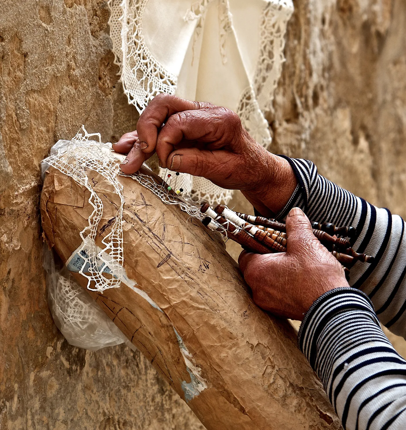 Two weathered hands working over a cylindrical lace pillow, throwing pairs of wooden bobbins across a partly finished strip of lace pinned to a paper pattern