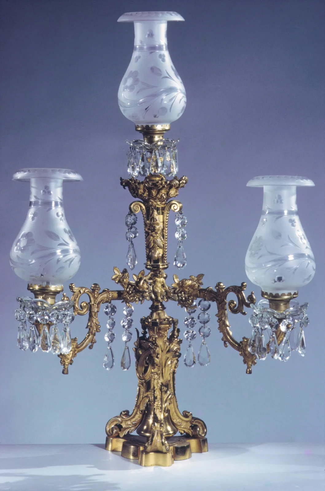 Three burner gas lamp of about 1855 in gilt bronze, the central burner raised above two curved side arms, each carrying an etched glass shade, with cut glass prism drops hung beneath and a shaped base