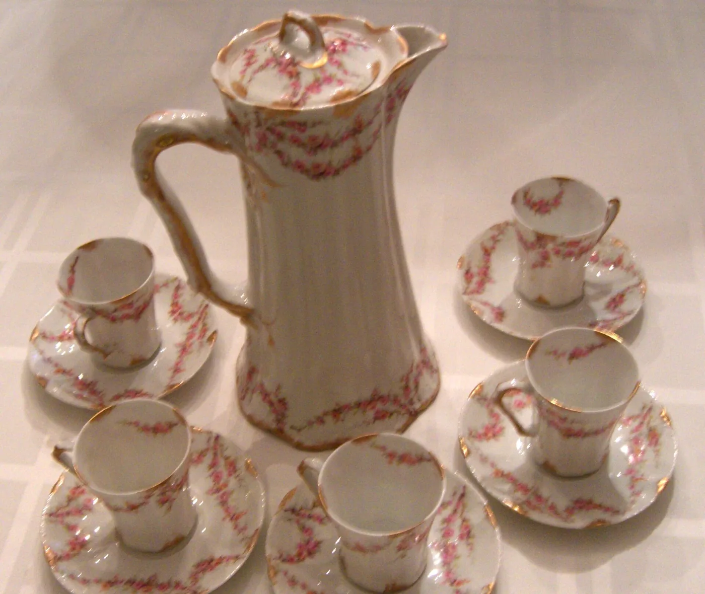 Tall tapering porcelain chocolate pot with a lid and long handle, surrounded by six matching cups and saucers decorated with pink rose garlands and gold