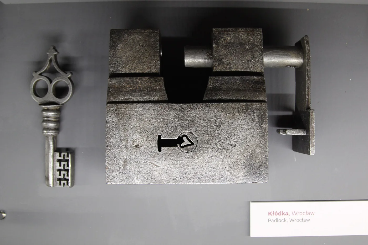 Large rectangular iron padlock with a heavy shackle mounted on a grey museum panel, its matching iron key with a pierced bit displayed beside it