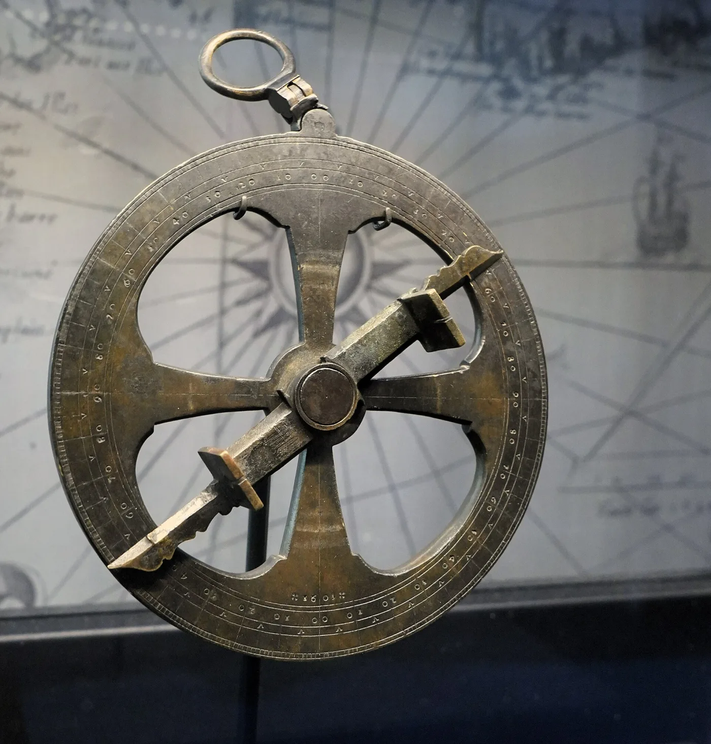 Heavy brass disc pierced with four large openings, a graduated rim and a pivoting alidade with two sighting vanes across its centre, suspended from a ring above a printed map