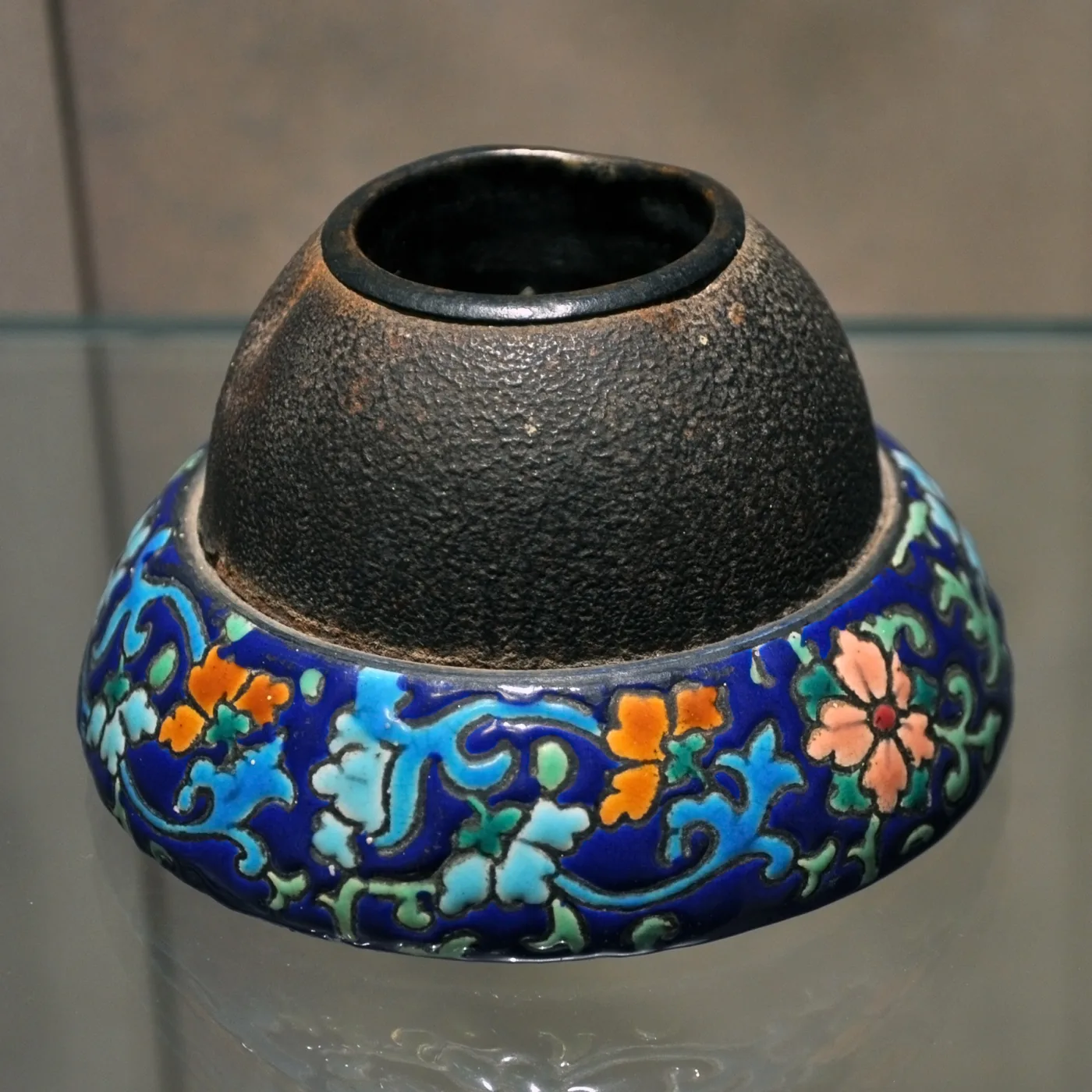 Squat match striker with a deep cobalt enamelled base decorated with turquoise, orange and green flowers, surmounted by a dark, coarsely textured metal dome with a hole at the top