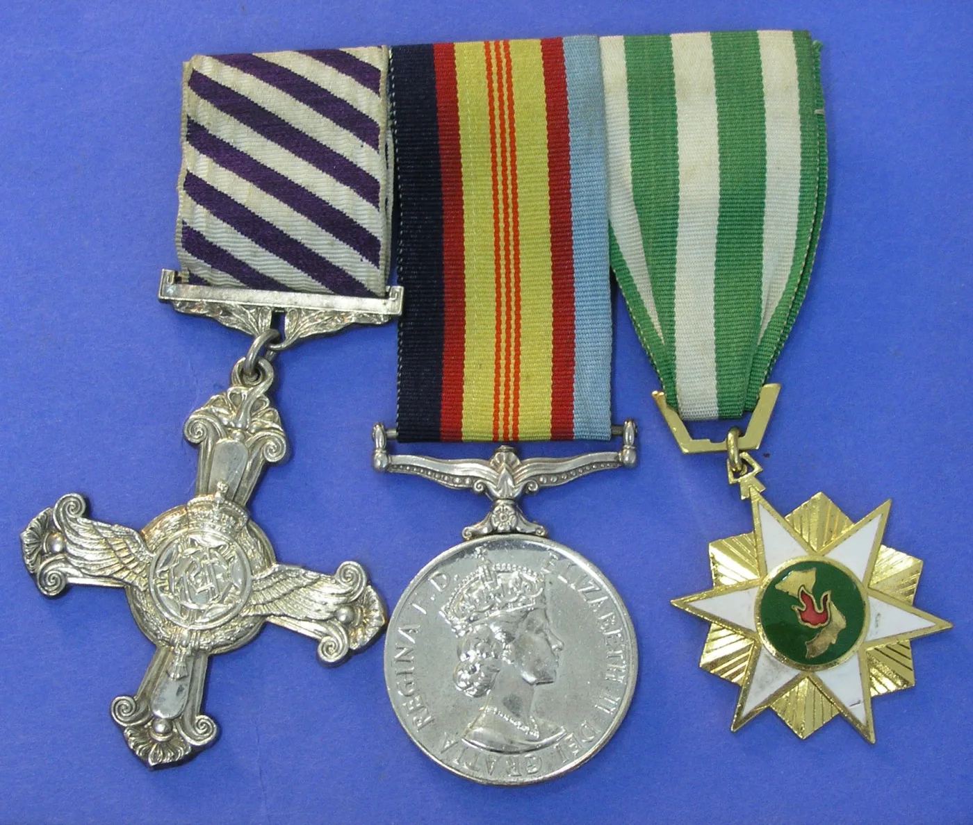Three medals laid side by side on a blue ground: a silver flying cross on a striped purple and white ribbon, a round silver medal with a crowned young queen's head, and a gold six-pointed star with a green and white enamelled centre