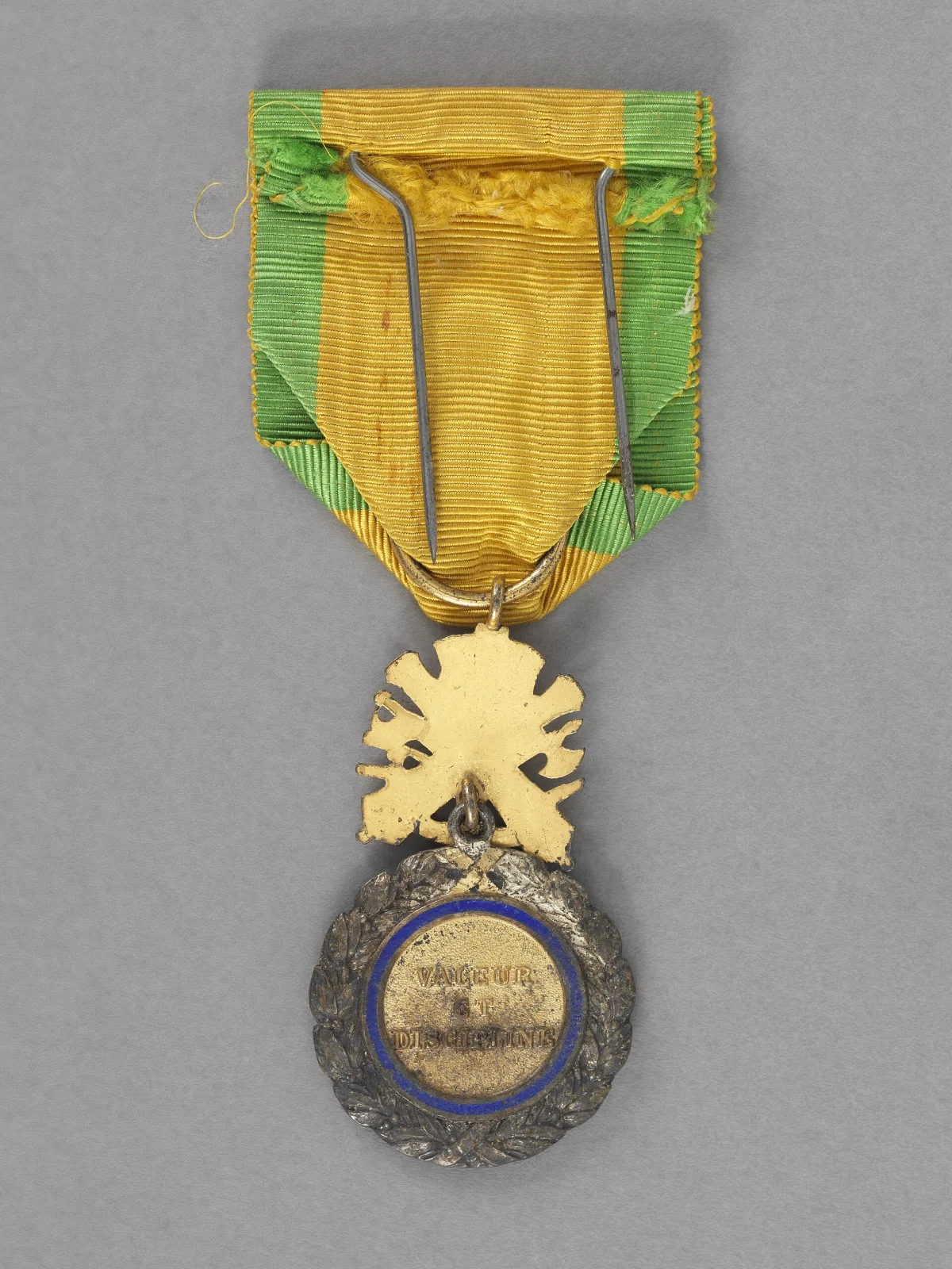 French Medaille militaire seen from behind, its yellow ribbon with green edges threaded over a metal suspension and the reverse of the enamelled medal below, on a grey ground