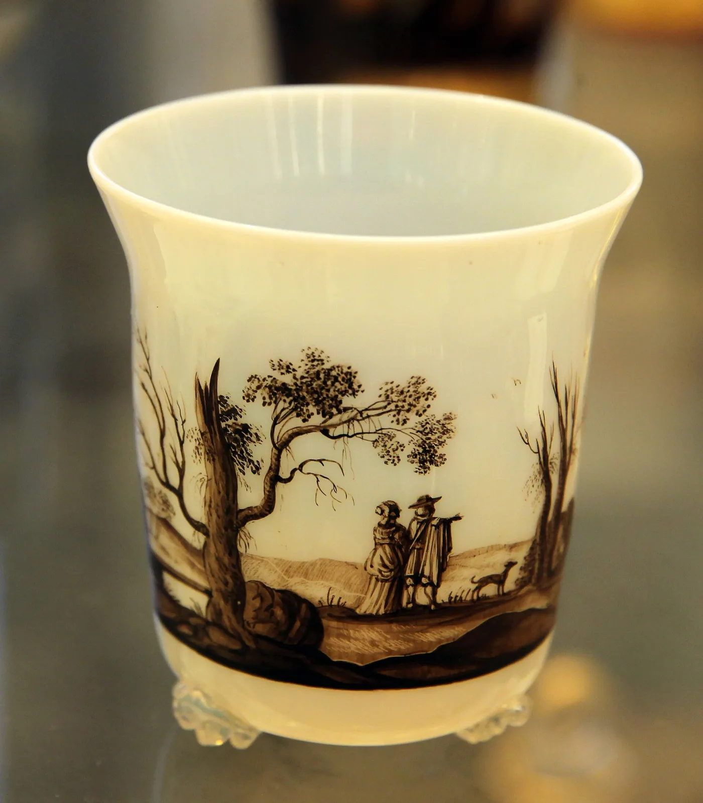 Cream-white glass beaker painted in brown-black enamel with two figures and a dog in a bare landscape