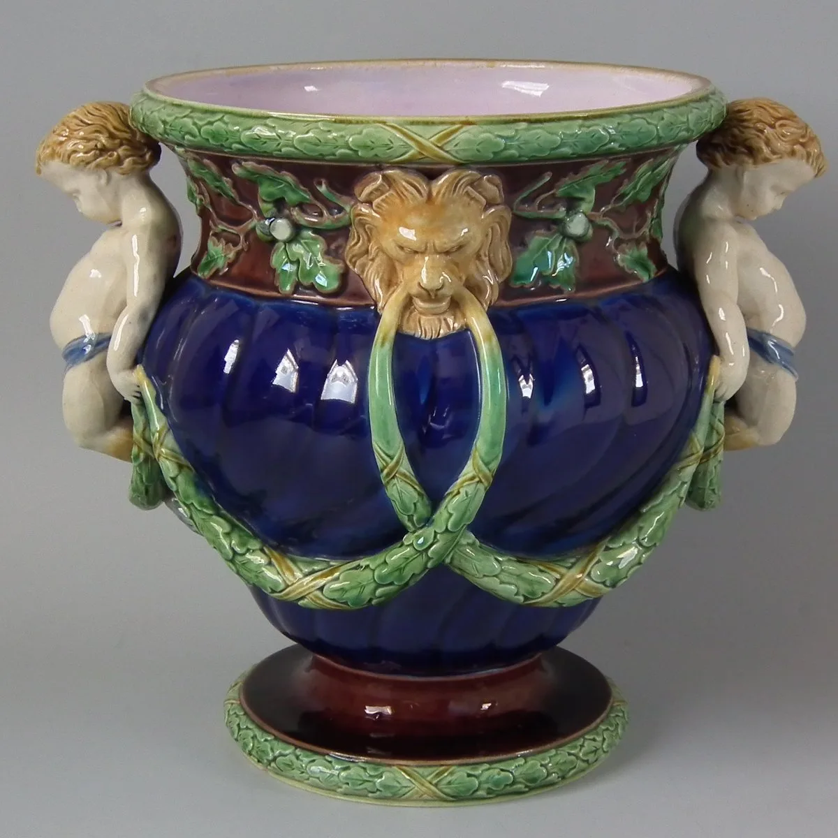 Deep cobalt-blue lobed jardinière with green oak-leaf and acorn garlands at the rim and swagged across the body, a tawny lion mask holding a ring at the front, and a pale modelled infant figure at each side, on a dark red foot