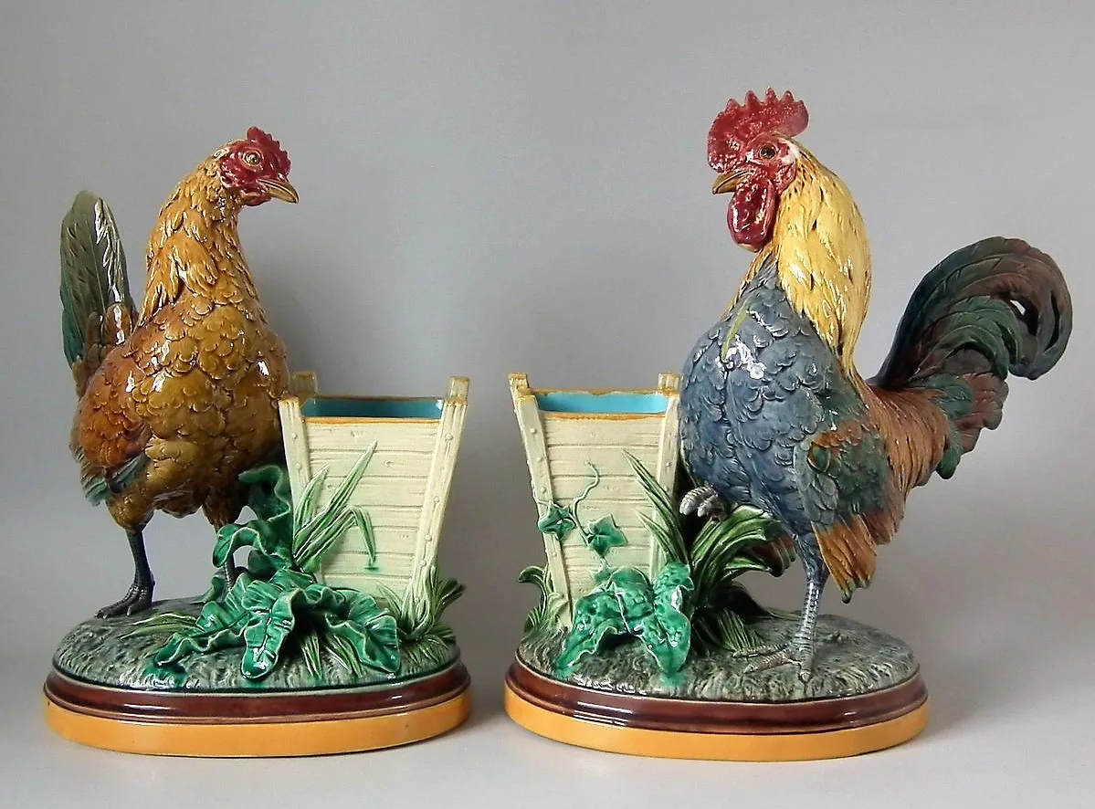 A pair of majolica birds, a hen and a cockerel with brown, grey and green plumage, each standing beside a woven basket on an oval green and ochre base