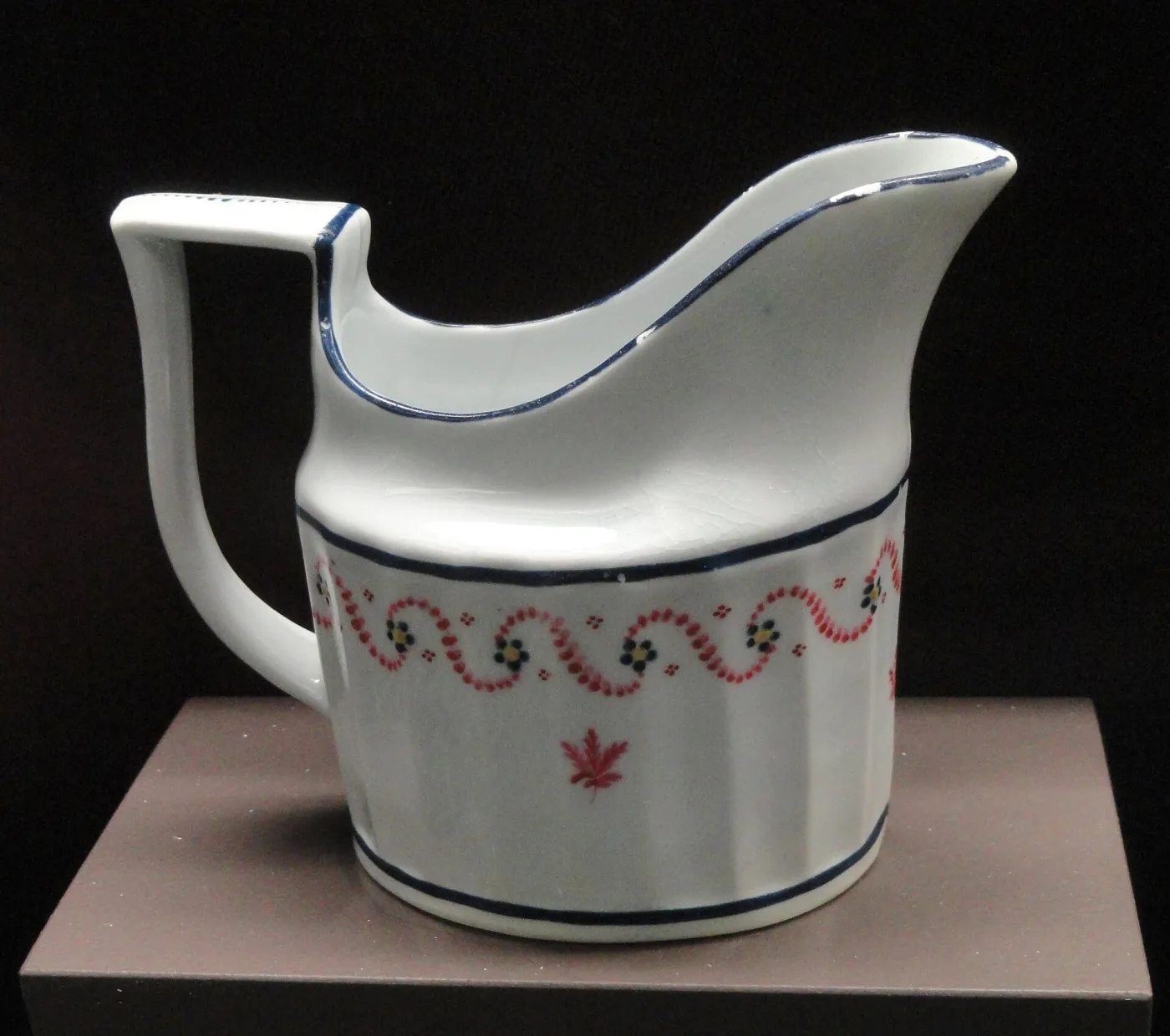 Small white fluted jug of oval section with a high looping handle, decorated with a thin blue line at the rim and a delicate pink and green swag around the body