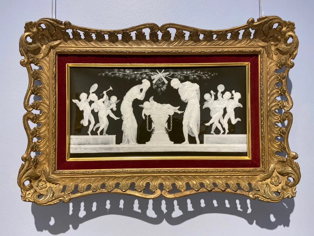 Rectangular plaque in an ornate gilt frame, its deep brown ground carrying a scene of pale translucent white figures dancing and playing music, built up in low relief