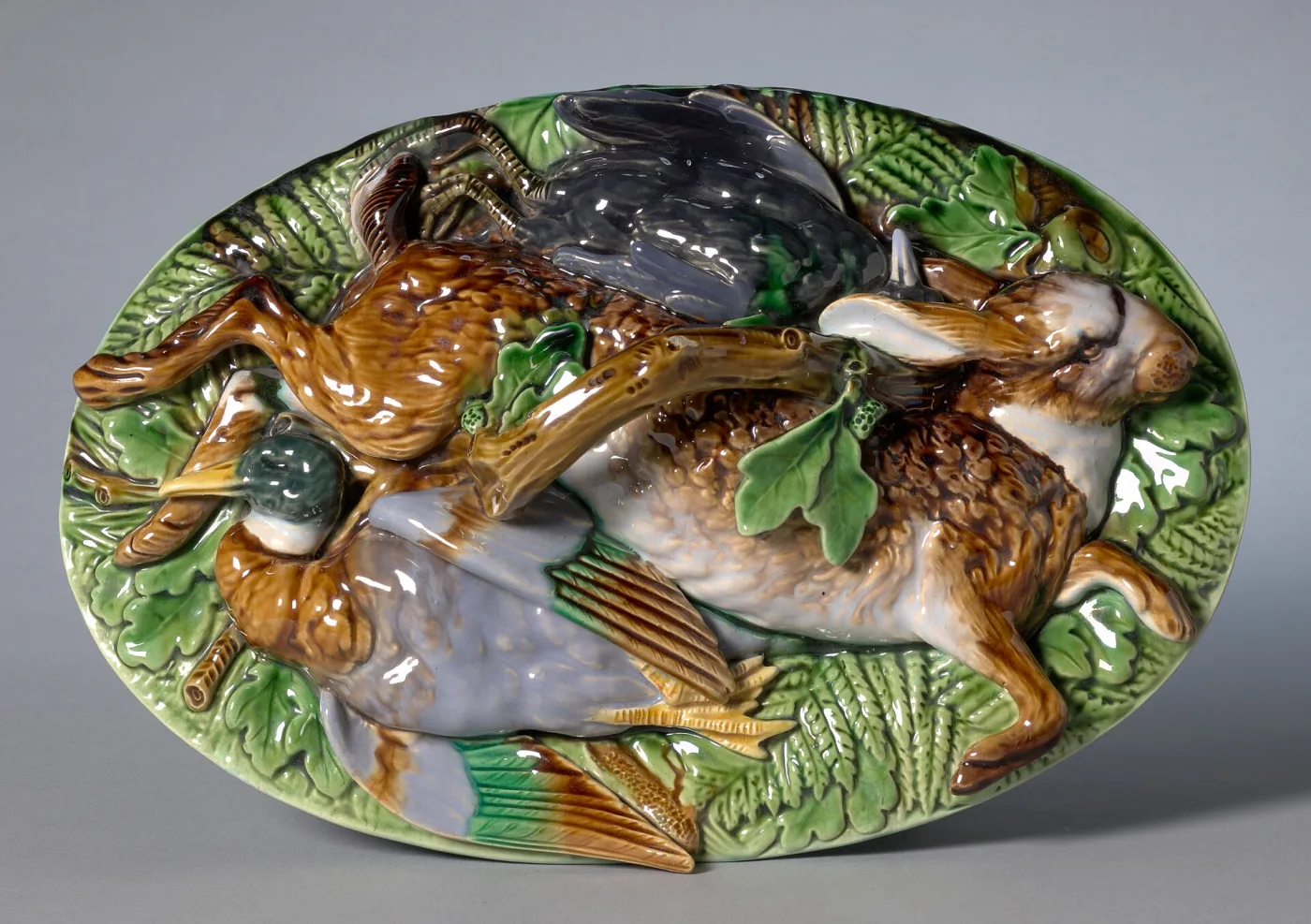 Oval majolica dish whose cover is modelled as a heap of dead game — a hare, a duck and smaller birds — lying among green leaves, the whole under rich brown, grey and green glazes
