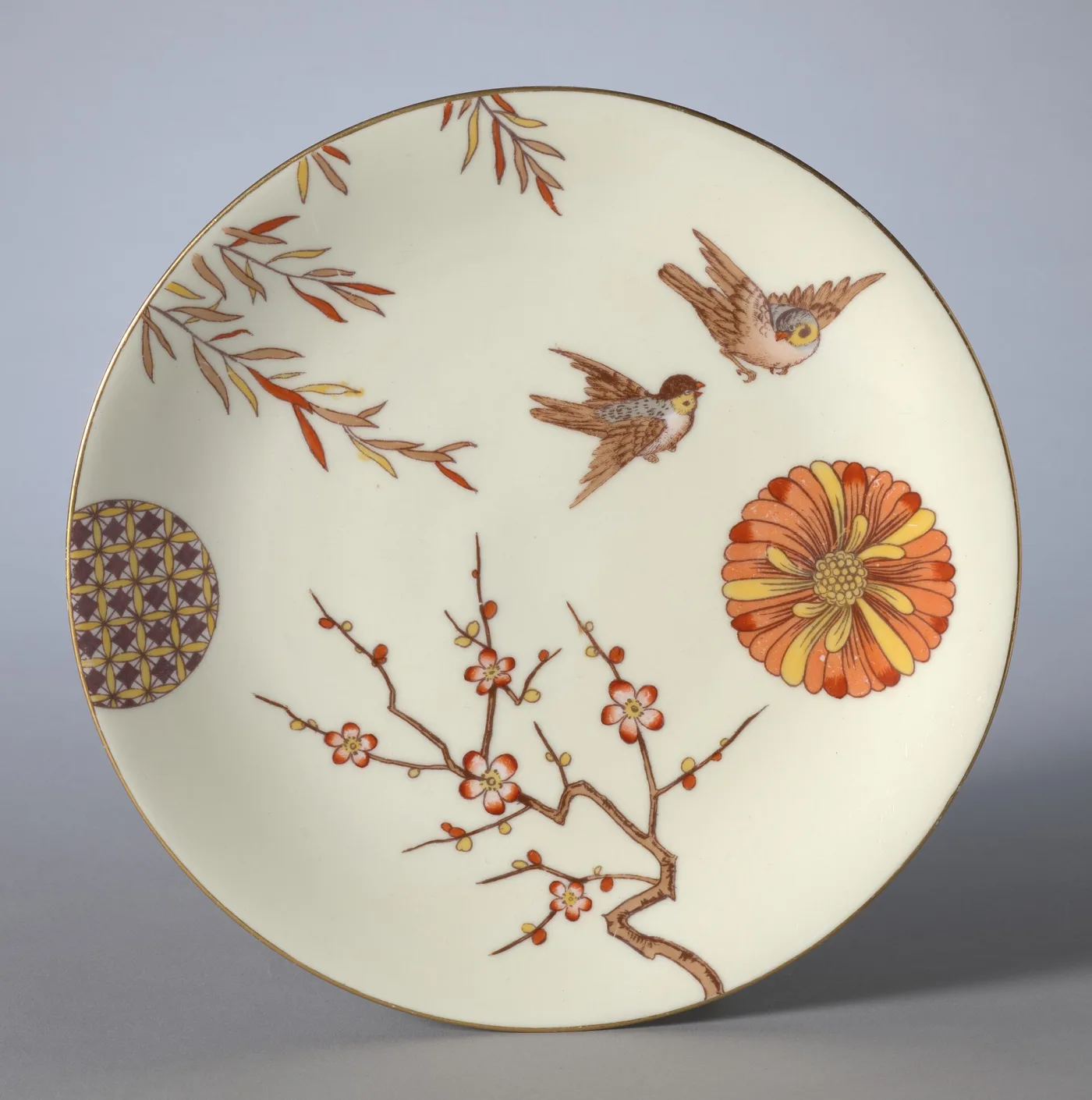 Cream porcelain plate decorated asymmetrically in soft red and brown with a flying bird, a branch of prunus blossom, a stylised chrysanthemum disc and a panel of geometric diaper