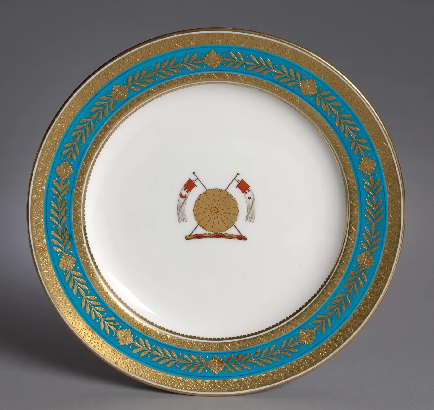 White porcelain plate with a broad turquoise border overlaid in gold with a formal leaf pattern, a narrow gilt rim, and a small painted crest of crossed instruments in the centre