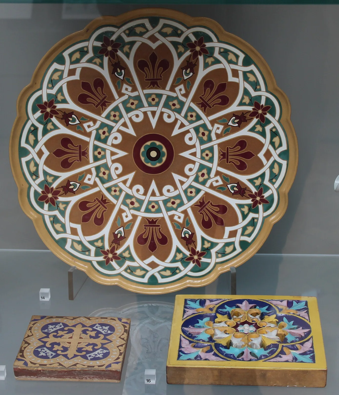Museum case holding a large scallop-edged circular tray decorated with a radiating pattern of brown, cream, green and dark red compartments, with two square tiles in raised mounts displayed flat in front of it